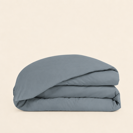 A folded dusty blue bedding item, likely a duvet cover or comforter, stacked neatly on a solid, warm off-white background.