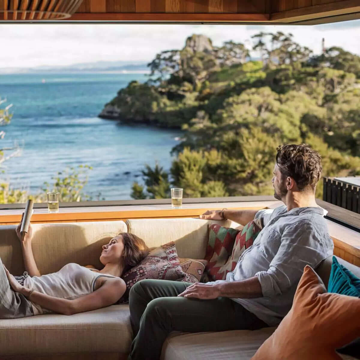 A couple sitting on a sofa inside a hilltop home, gazing out over the ocean through large windows.