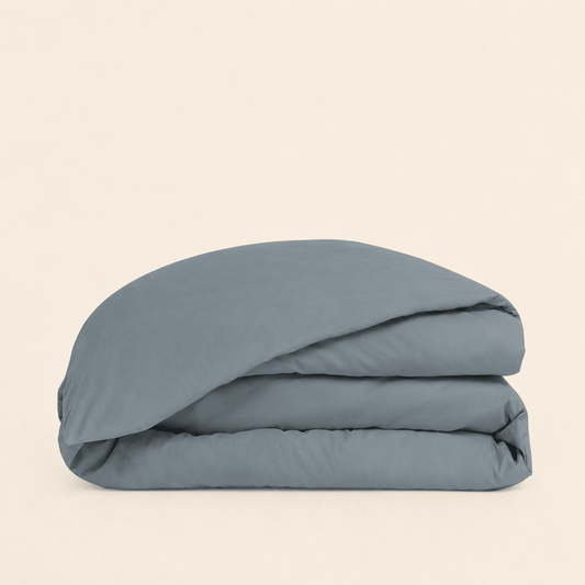 A folded dusty blue bedding item, likely a duvet cover or comforter, stacked neatly on a solid, warm off-white background.