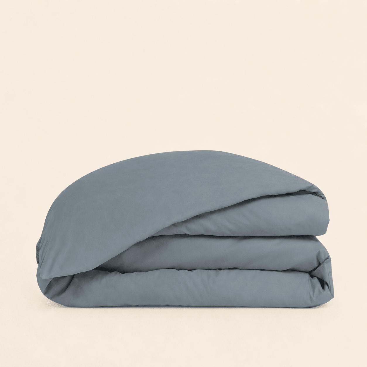 A folded dusty blue bedding item, likely a duvet cover or comforter, stacked neatly on a solid, warm off-white background.