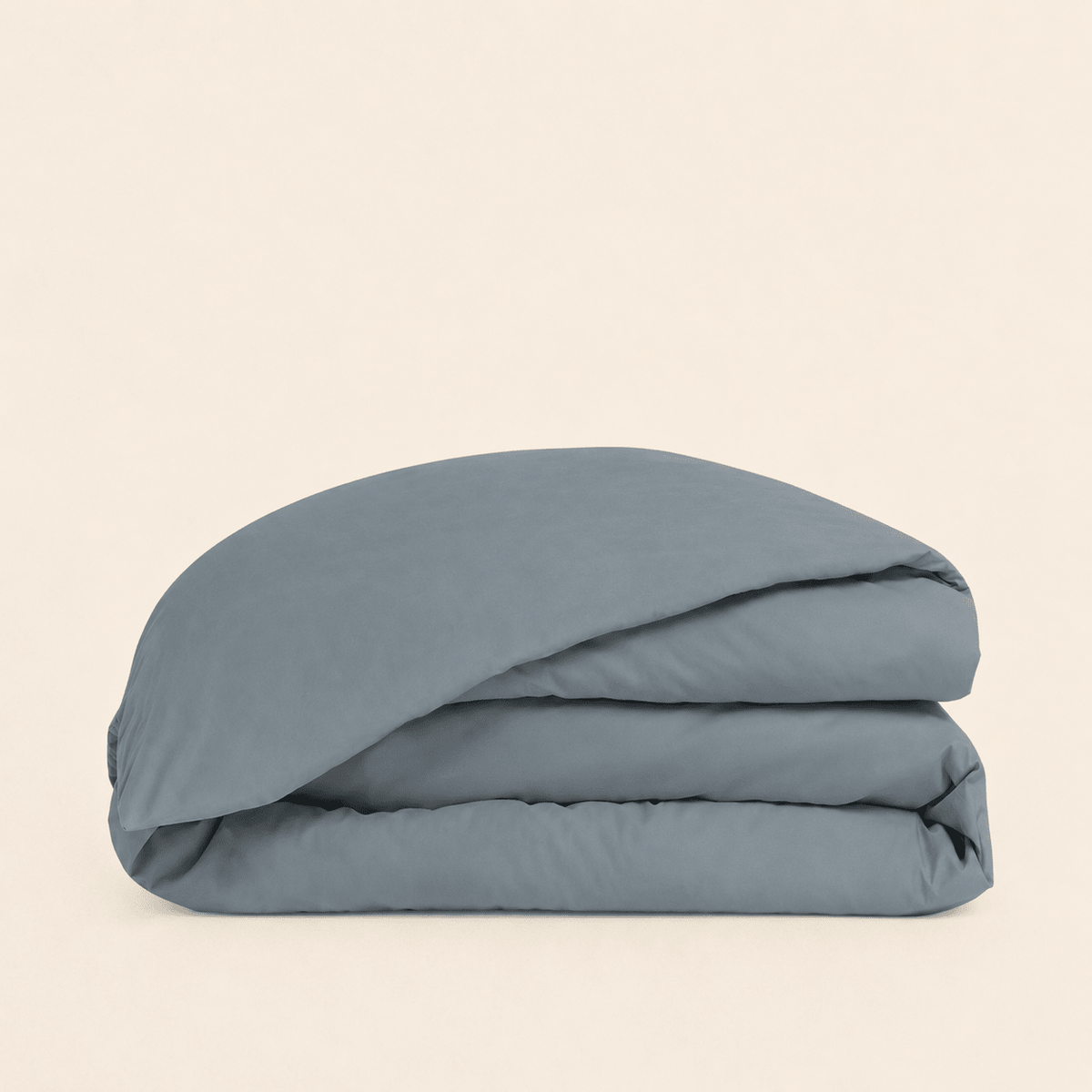 A folded dusty blue bedding item, likely a duvet cover or comforter, stacked neatly on a solid, warm off-white background.