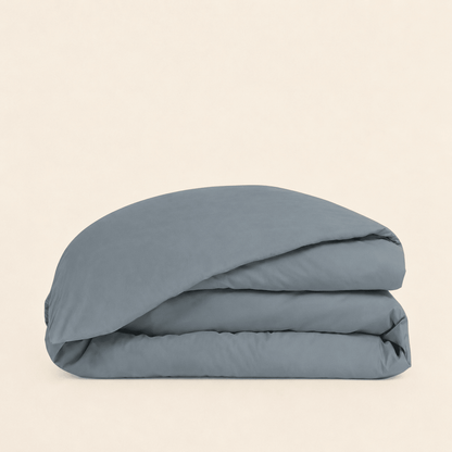 A folded dusty blue bedding item, likely a duvet cover or comforter, stacked neatly on a solid, warm off-white background.